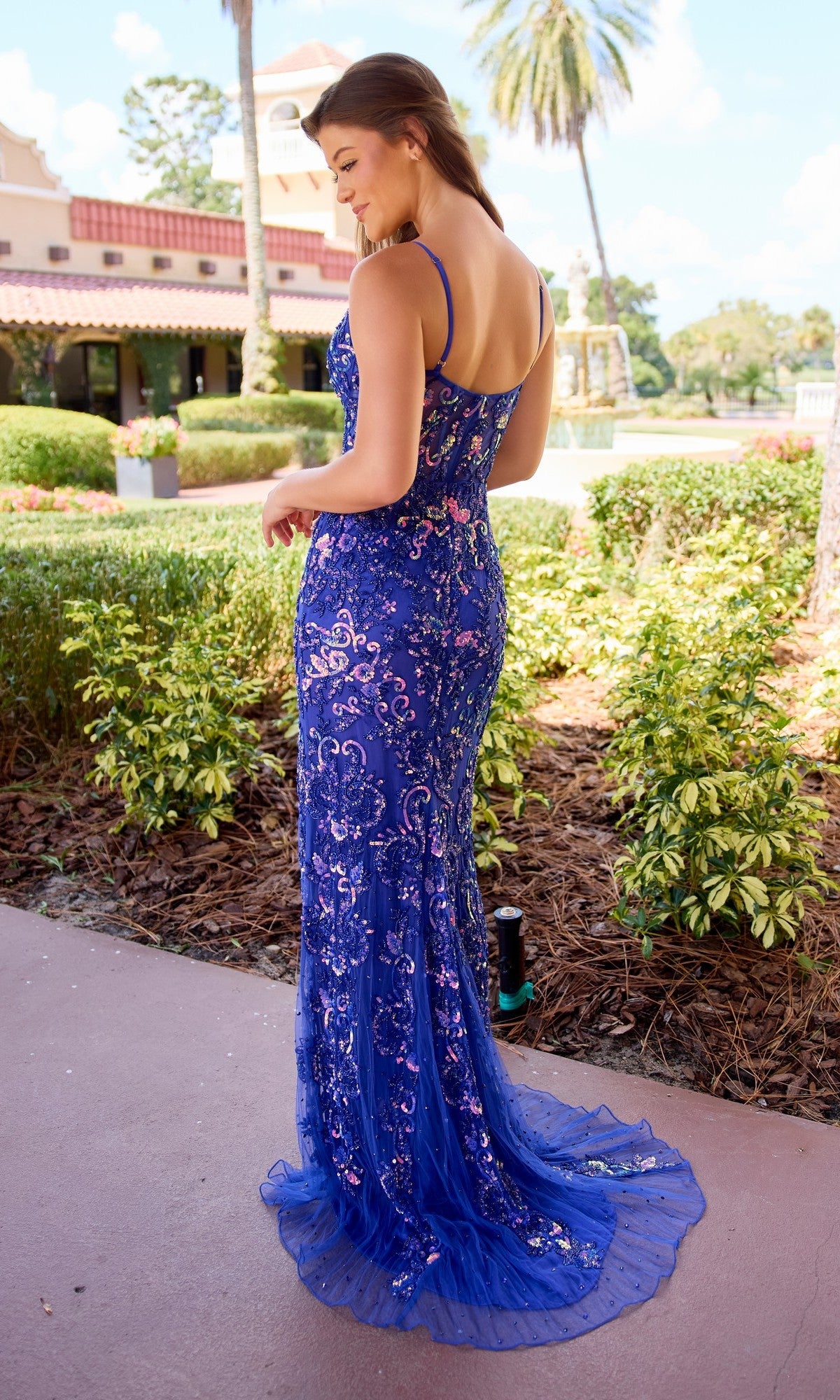 A woman stands outdoors in the Long Prom Dress: Primavera 4549, a floor-length blue sequin fitted sheath gown with thin straps, surrounded by greenery and buildings.